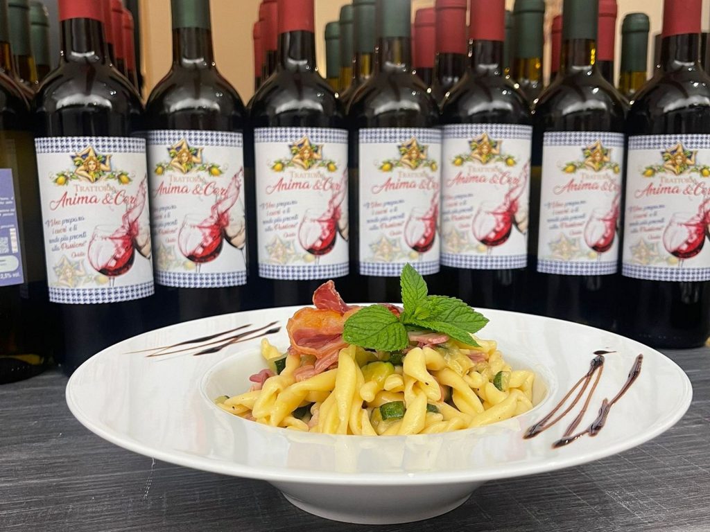 Bottiglie di vino rosso siciliano con un piatto di pasta fresco in esposizione alla Trattoria Anima e Cori